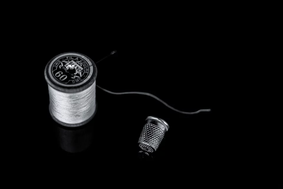 Photographie en noir et blanc d’une bobine de fil et d’un dé à coudre posés sur un fond noir, éclairés de façon à faire ressortir les textures du métal et du fil.