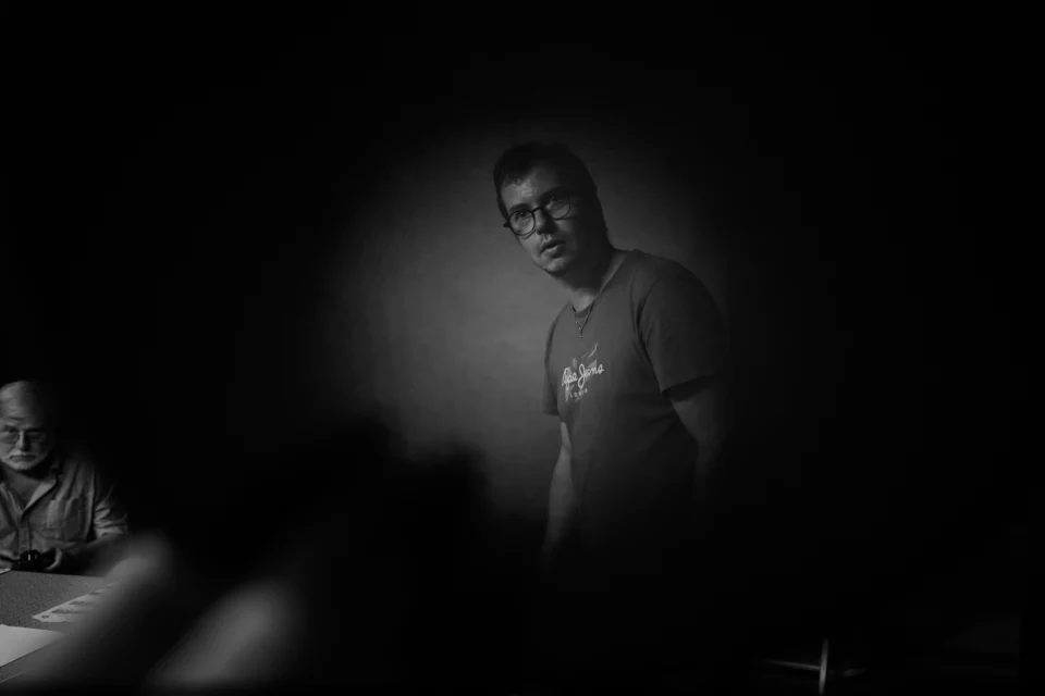 Photographie noir et blanc prise à travers un nid d’abeille de flash. Nicolas se tient au centre, éclairé dans la pénombre, tandis que Michel est assis à gauche. Les alvéoles floues de l’accessoire forment un cadre sombre au premier plan.