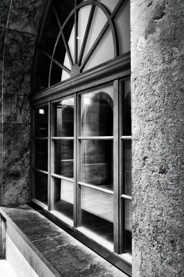 Fenêtre cintrée en bois photographiée en noir et blanc, encadrée par des murs de pierre et parcourue de reflets dans les vitres.