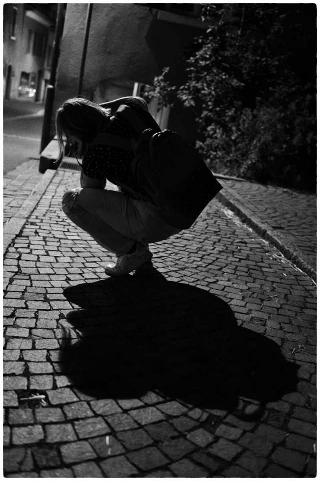 Personne accroupie sur une rue pavée photographiée en noir et blanc, avec une grande ombre projetée devant elle.